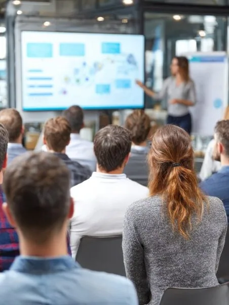A group of people seated facing a presenter who is pointing at a large presentation screen in a bright, modern conference room.