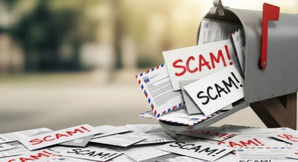 A mailbox overflowing with letters labeled "SCAM!" surrounded by scattered envelopes and papers on the ground.
